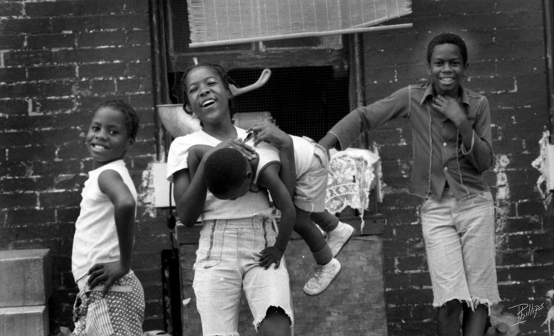 A photograph of four children laughing and playing. They have brown skin and are smiling and posing in front of the camera. 