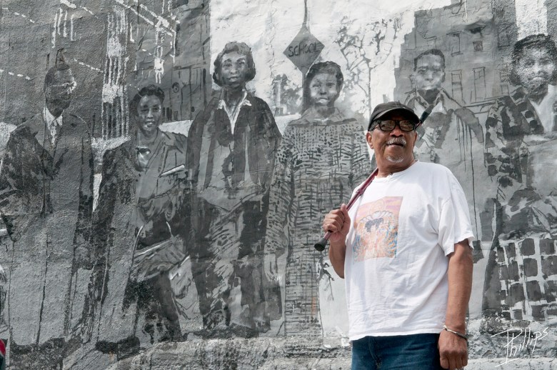 Irving Henry Webster Phillips Jr. standing in front of a mural. He is a brown skinned man wearing a white tshit, black baseball cap, and black thick rimmed glasses. 