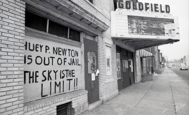 A black and white scan of a derelict theater. A hand painted sign reads:  Huey P. Newton Is Out Of Jail. The Sky Is The Limit" 