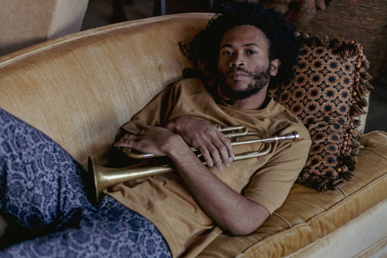 A man with brown skin lays on a couch holding a trumpet. 
