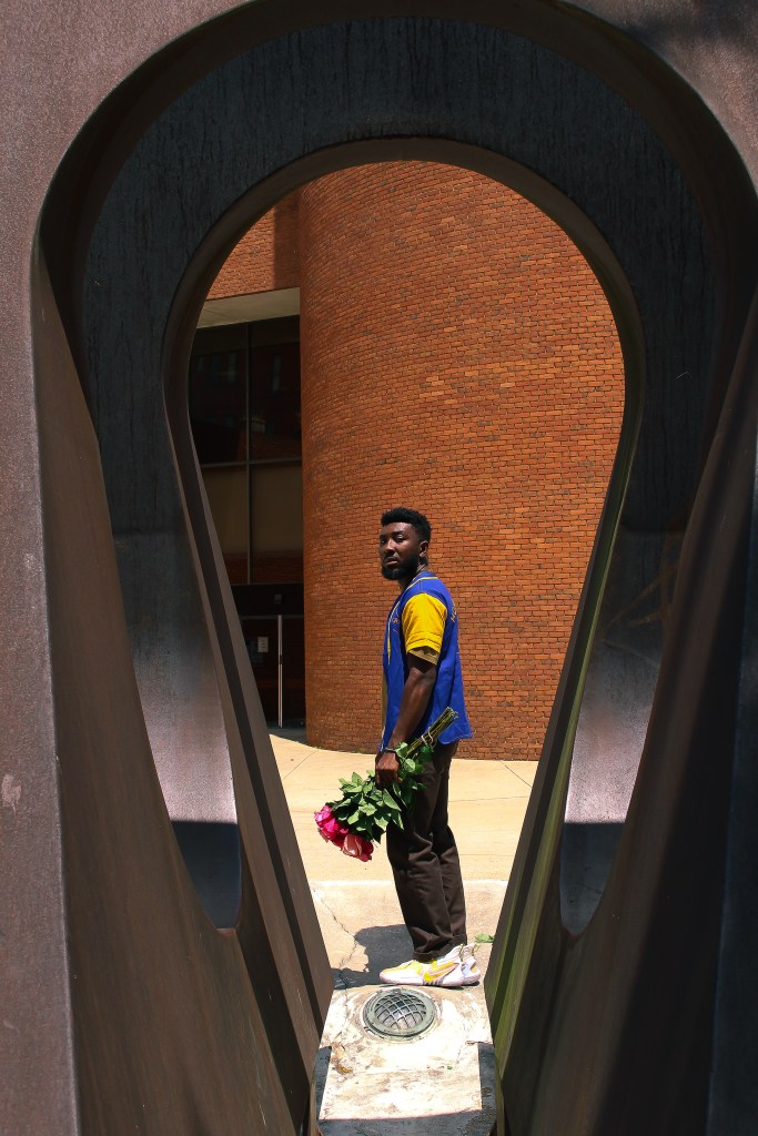 A man with brown skin is posing holding a bouquet of flowers behind a scultpure. 