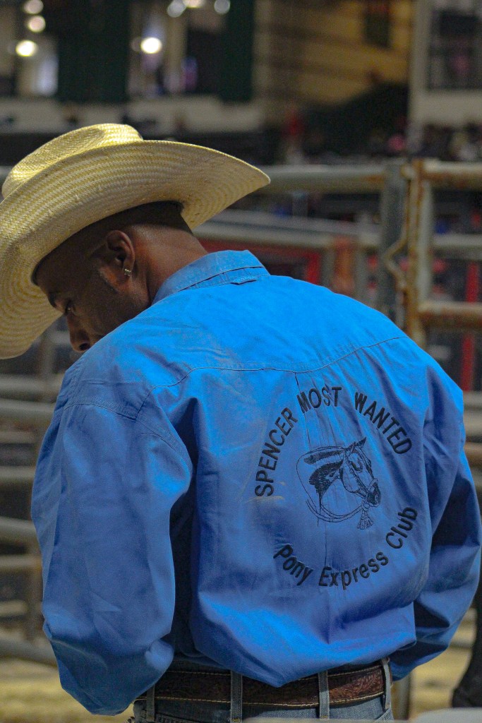 A competitor from Oklahoma’s Spencer Most Wanted Pony Express Club in the 35th Annual Bill Pickett Invitational Rodeo. He wears a blue long sleeve t-shirt, blue jeans, and a tan cowboy hat. 
