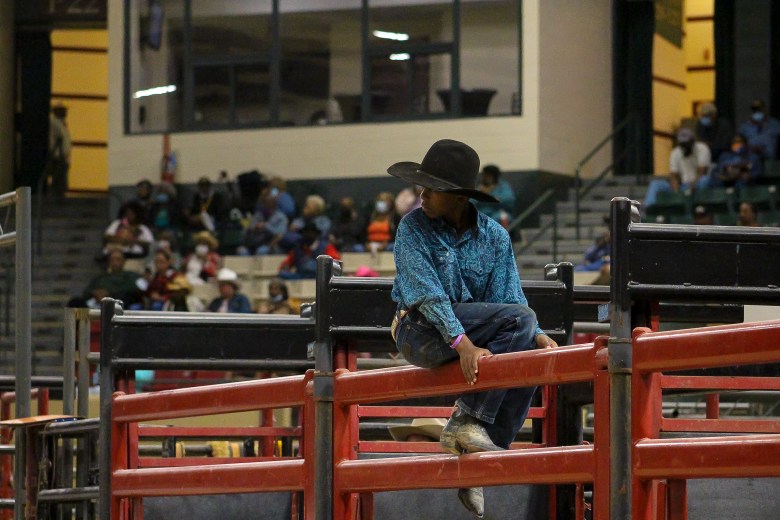 A young cowboy surveys the rodeo. He is sitting on a red metal gate, wearing a blue shirt and blue jeans, and a black cowboy hat. 