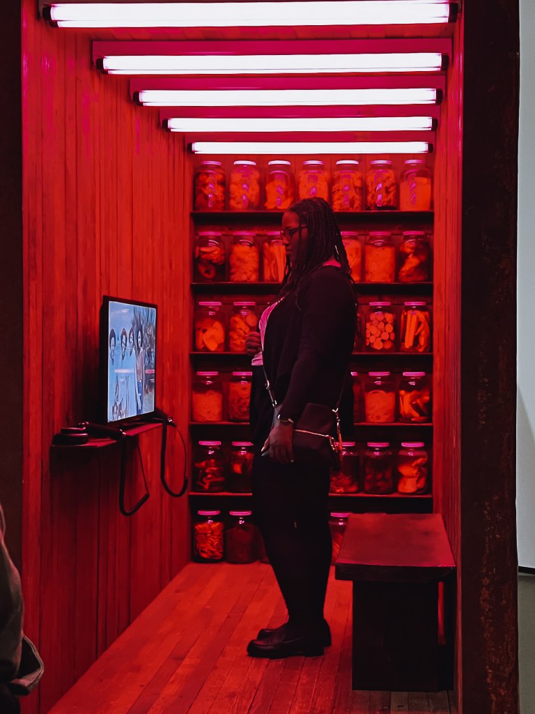 A visitor to the BMA looks at a video work at the Baltimore Museum of Art, they are standing and behind them are rows of canned foods. 