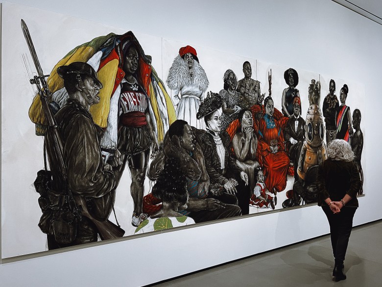 A visitor in a gallery looking at a large drawing of Black figures. They are wearing various costumes. 