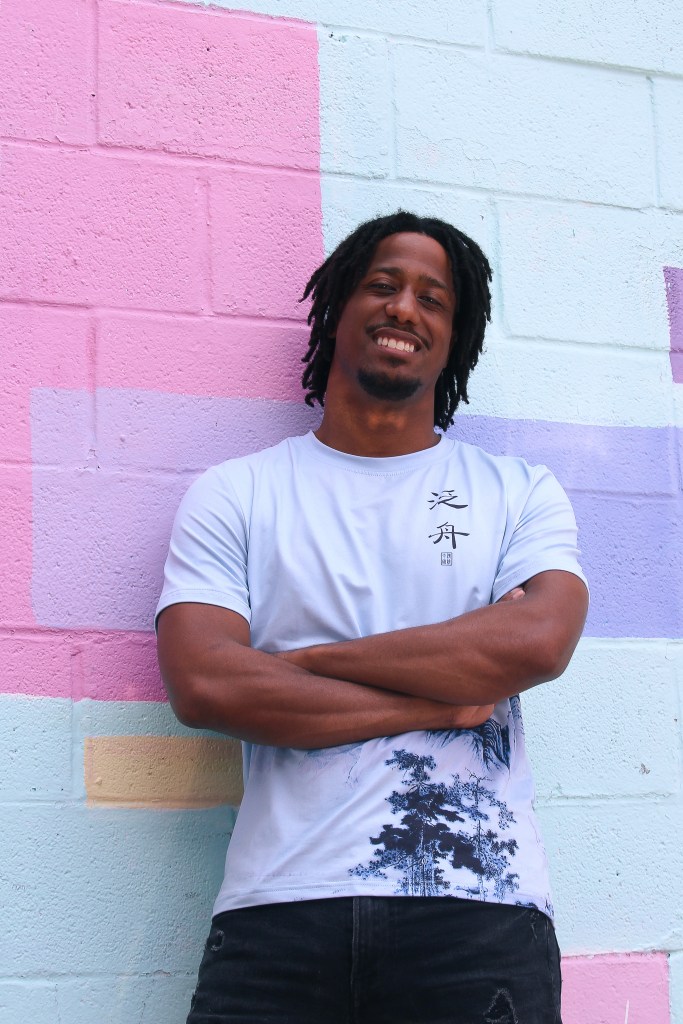 A man with brown skin stands in front of a painted wall. 