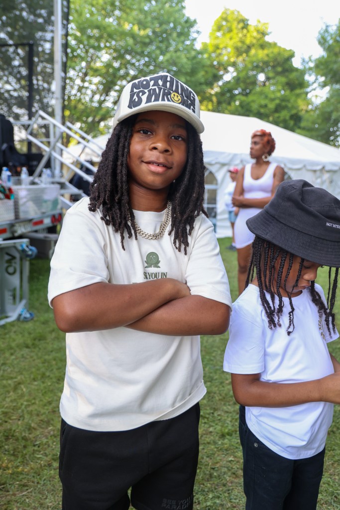 A young person smiles for the camera with their arms folded in front of them. They wear a cap that says "nothing is wrong." Another young person wearing a black bucket hat, a white t-shirt, and black shorts looks down.