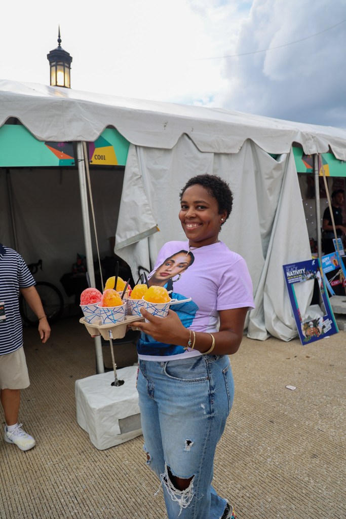 A person carries a tray of snowballs. She is wearing a tshirt that has the artist Sade on it.. 