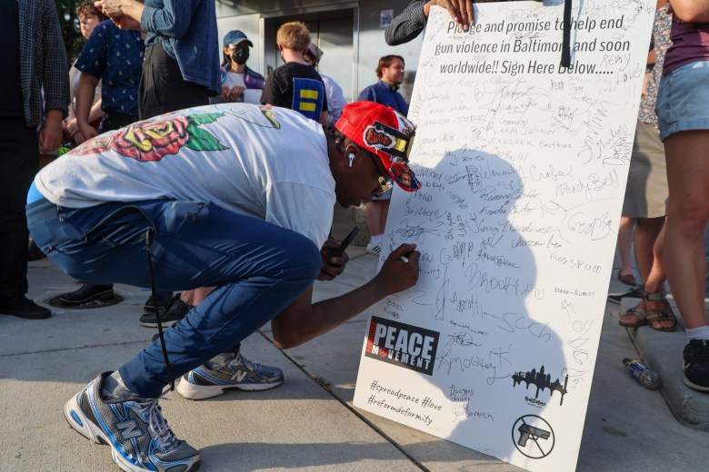 A person signs a large white board. It reads: "pledge and promise to help end gun violence in Baltimore and soon worldwide!! Sign here below..." The sign is covered in signatures. 