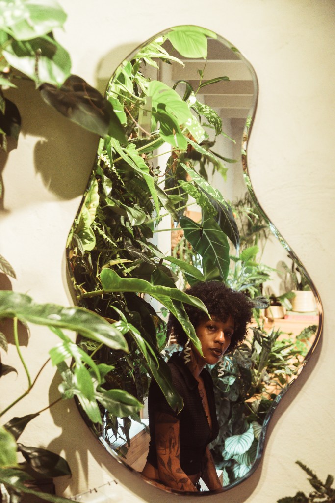 Photograph of a woman with brown skin sitting surrounded by green plants. She is looking at her reflection in a wavy shaped mirror. She wears a black top, her hair is dark, she is wearing dark lipstick and large decorative earrings. 