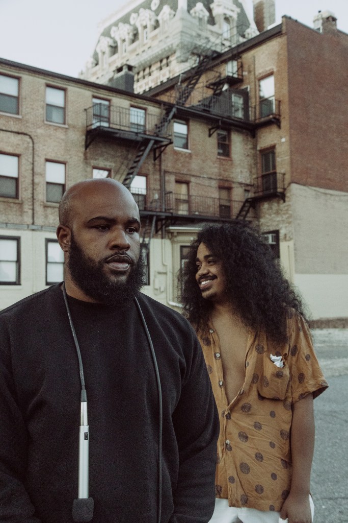 Two men are pictured, both have brown skin, one is bald and wearing a black long sleeve shirt with a microphone around his neck, the other has curly hair and is wearing a brown shirt with brown details, he is smiling. 