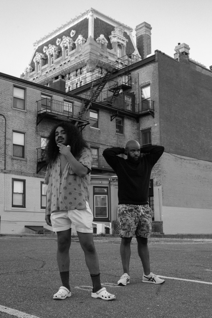Black and white photograph of two men with brown skin. One is wearing a black long-sleeved shirt and has a bald head, the other figure is wearing a polka dot shirt and he has long curly hair. They are standing in front of brick buildings. 