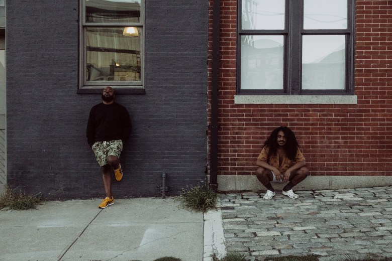 Two men are pictured in front of a dark wall and a red brick wall. One is crouching and looking at the camera, he has curly hair, the other is bald and is standing with one leg rested against the dark wall. 