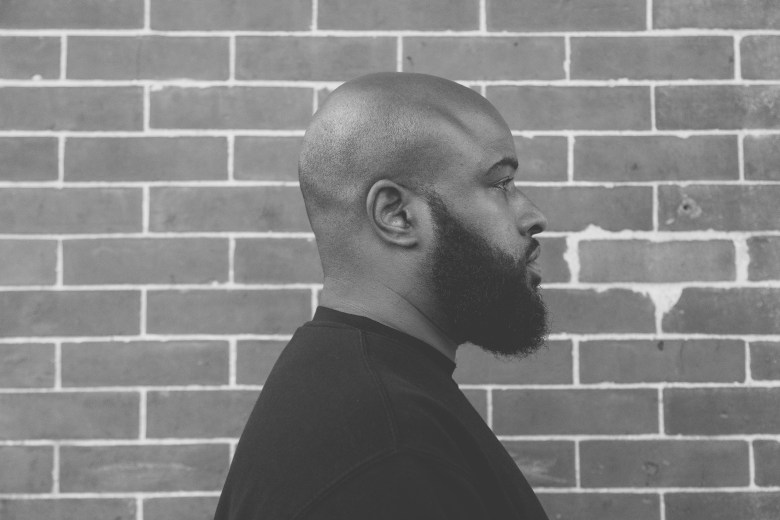 A black man with a bald head is facing to the right of the image. He has a beard and is wearing a black shirt and he is standing in front of a brick wall. 