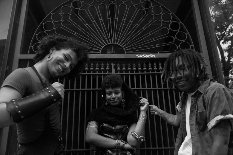A black in white photo with three members of BLKVAPOR. They are pictured in front of an iron gate. Jamie Proxy Grace is on the far left, wearing hoop earrings with curly brown hair and a leather sleeve, in the middle Safra Tadesse weas a black collar and is smiling, wearing shiny earrings, and to the right Kirby Bell is pictured, Bell is pictured wearing glasses and has dreadlocks. 
