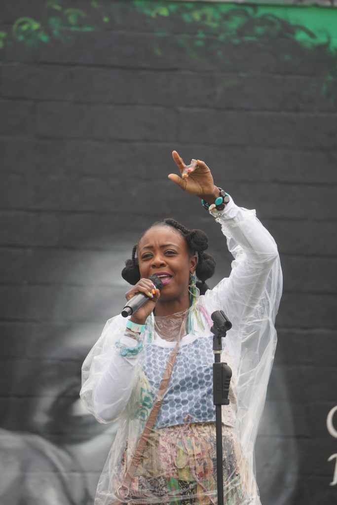 A woman with brown skin speaks into a microphone. She has her arm raised. 