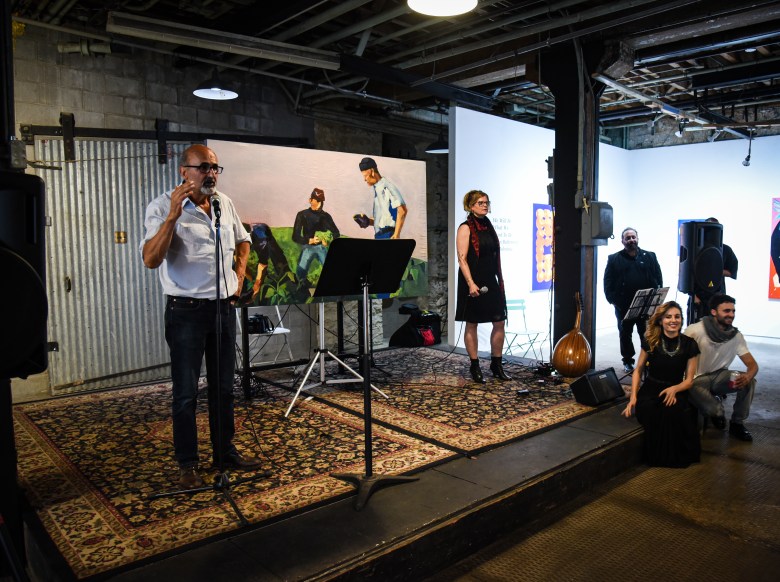 This is a photo of a group of people presenting on a stage. A male figure is gesturing and speaking to an audience. 