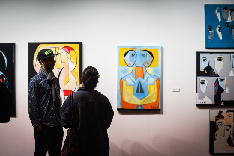 Two people talk in front of a wall of paintings.. 