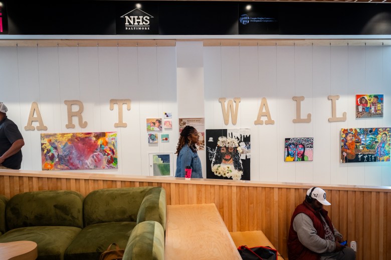 A woman walks past a white wall. The words "art wall" are spelled out on the wall with large letters. Brightly colored art is hanging on the  wall. 
