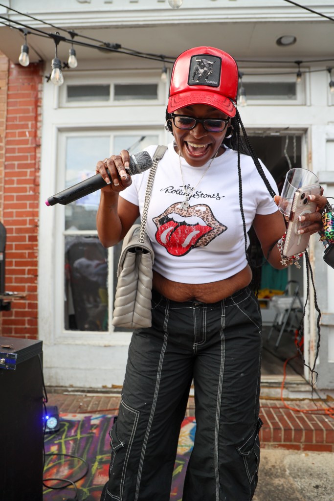 A person wearing a red cap, white top, and black pants speaks into a microphone. 