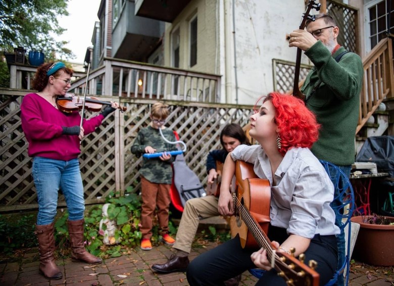 A family stands outside of a home, listening to a woman play a guitar. 
