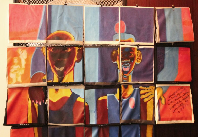 A color photo of an artwork. It is a series of T-shirts that resemble tiles, the shirts displayed together to make an image of two young boys with brown skin. Behind them, a figure is standing with its face obscured. The image is made of red, blue, purple, yellow, and white.